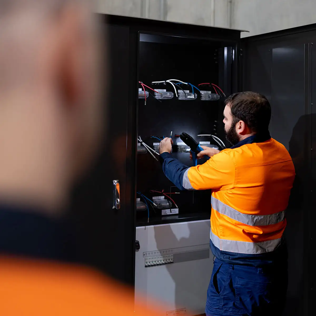 Electrician in high‑visibility gear inspecting and wiring an electrical switchboard during a service job
