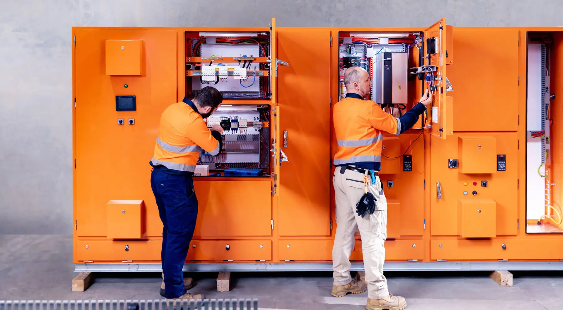 Electricians in high‑visibility gear inspecting and wiring an electrical switchboard during a service job